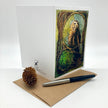 A white greeting card with an illustrated owl artwork on the front, displayed upright on a kraft paper envelope with a pen and a small pinecone prop on a clean white background.