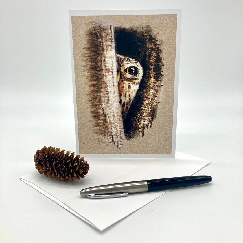 A vertical greeting card featuring a detailed illustration of an owl peeking through bark, displayed on a white surface with a pinecone and a black-and-silver pen in front of it.
