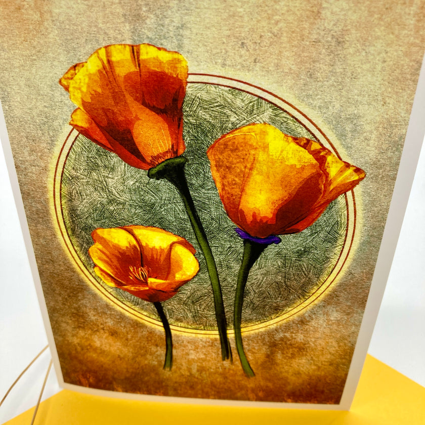 A close-up of a printed card or art print featuring three stylized orange-yellow flowers with green stems on a textured beige and brown background, photographed against a white surface with a yellow object at the bottom edge.