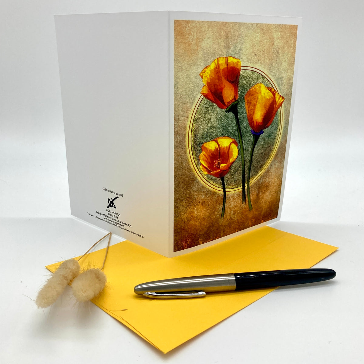 A white greeting card with a vintage-style illustration of three orange flowers on the front, displayed next to a yellow envelope and a black and silver pen on a white background.