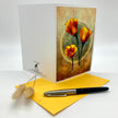 A white greeting card with a vintage-style illustration of three orange flowers on the front, displayed next to a yellow envelope and a black and silver pen on a white background.