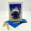 A vertical greeting card featuring a watercolor painting of a blue and gold teacup on a saucer, displayed standing upright on a blue envelope with a black and silver pen and two dried bunny tail stems on a white background.