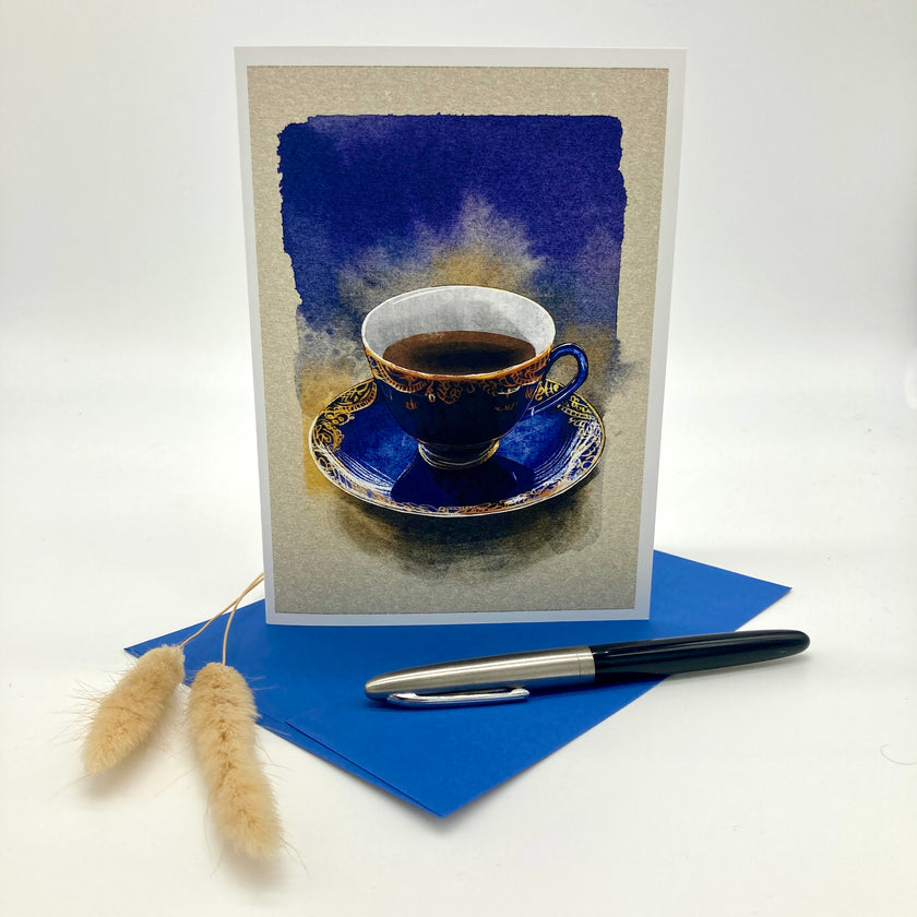 A vertical greeting card featuring a watercolor painting of a blue and gold teacup on a saucer, displayed standing upright on a blue envelope with a black and silver pen and two dried bunny tail stems on a white background.