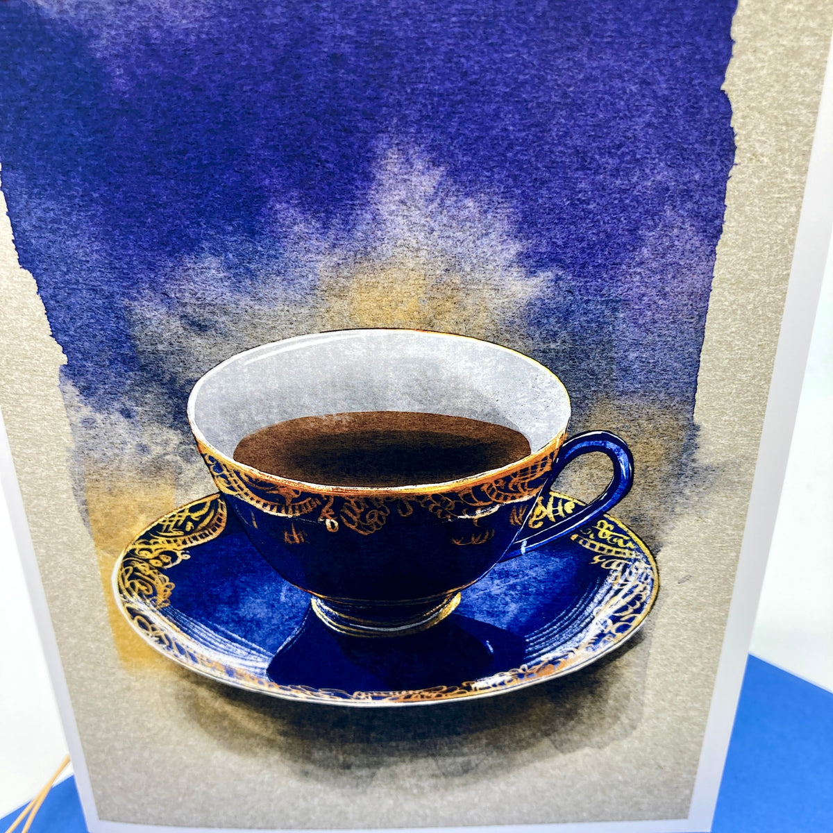 A card featuring a painted blue teacup and saucer with gold detailing, set against a textured blue and beige background.