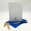 A white greeting card with black printed text stands upright on a blue envelope, with a black and silver pen resting on the envelope and dried beige grass stems to the left, photographed on a clean white studio background.