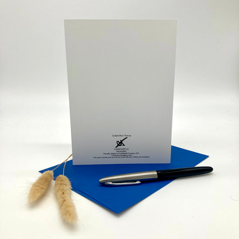 A white greeting card with black printed text stands upright on a blue envelope, with a black and silver pen resting on the envelope and dried beige grass stems to the left, photographed on a clean white studio background.