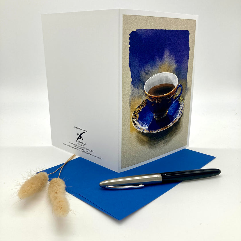 A white greeting card with an artistic print of a blue coffee cup on a saucer, displayed on a white background with a blue envelope, a black and silver pen, and two dried bunny tail stems.