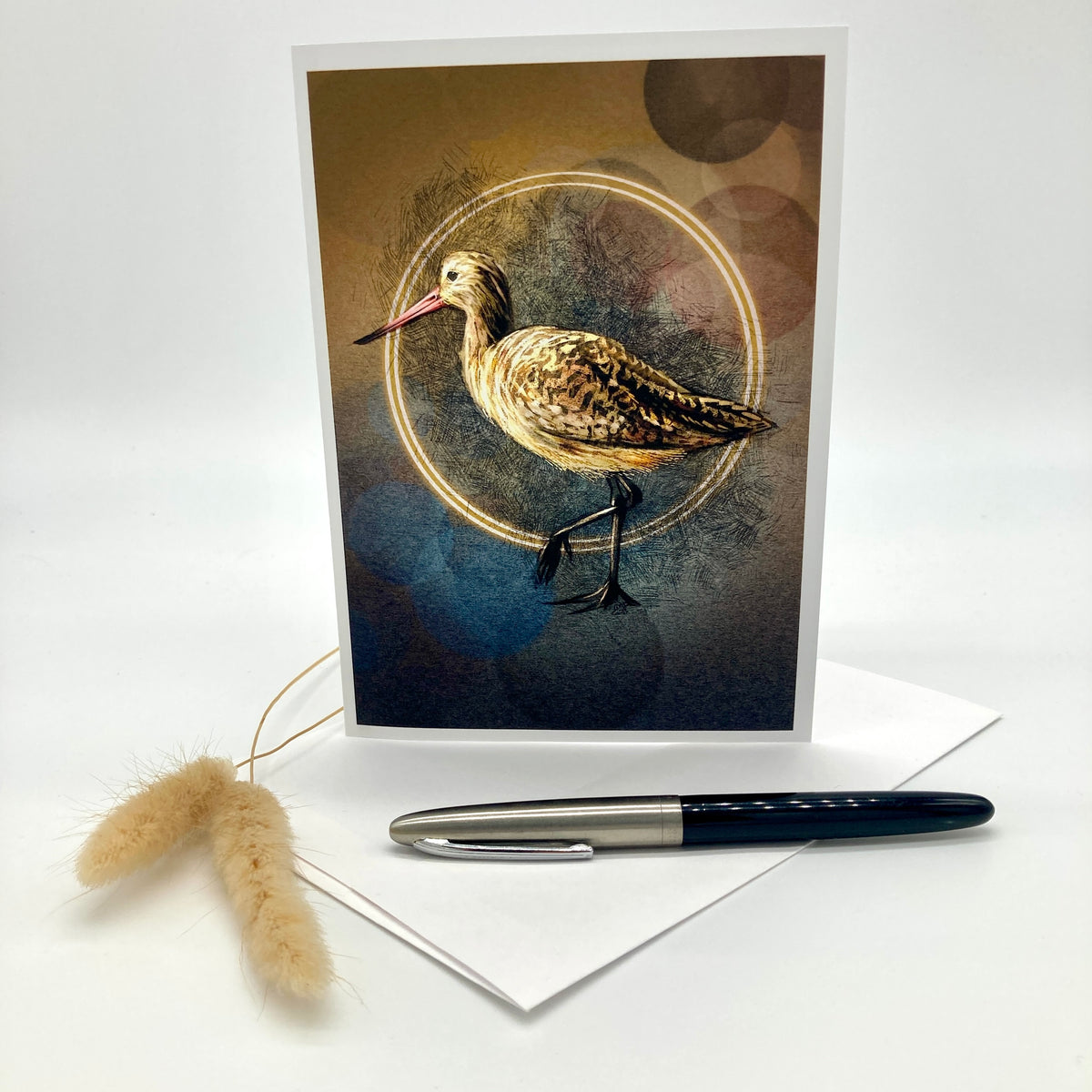 A vertical greeting card featuring a detailed illustration of a brown and beige bird standing within a glowing circular ring, set against a dark textured background. The card is displayed upright on a white surface with a white envelope, a black pen, and dried bunny tail grass as props.