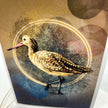 A close-up of an art print or card featuring a detailed illustration of a brown and beige bird with a pink beak, set against a textured background with gold circular lines and blue accents.