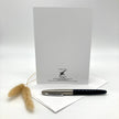A white stationery card with black printed text at the bottom stands upright on a white surface, with a black and silver pen resting on a white envelope in front and dried bunny tail grass to the left.