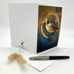 A white greeting card with an illustrated bird on the front, displayed upright with a pen and dried grass stems on a white surface. The card shows the Nature of Things brand logo on the inside.