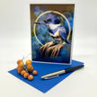 An illustrated greeting card featuring a Belted Kingfisher bird on a blue background with abstract glowing bubble orbs behind it. Posed with a sprig of yellow berries, an ink pen, and a dark blue envelope.