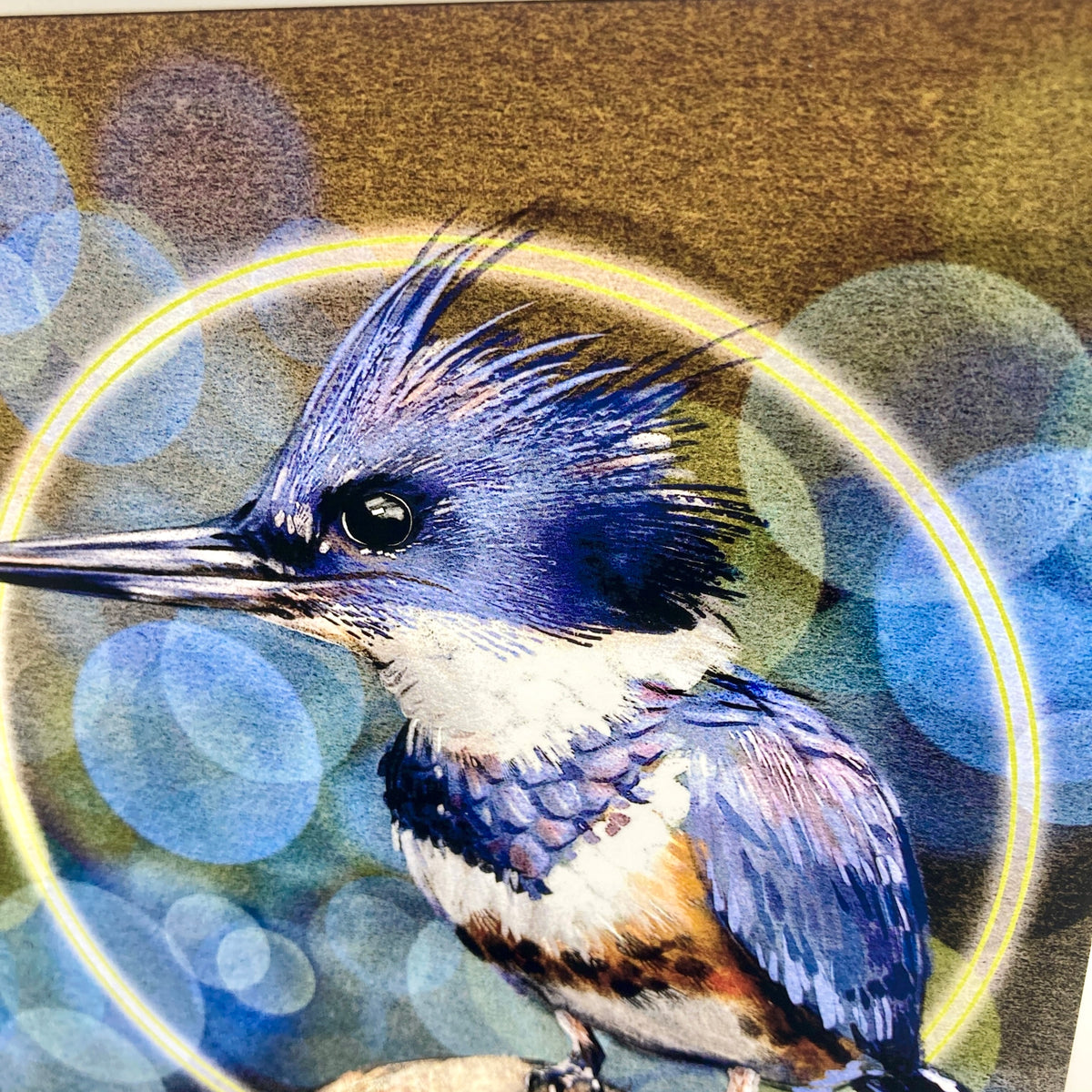 A close up of n illustrated greeting card featuring a Belted Kingfisher bird on a blue background with abstract glowing bubble orbs behind it. 
