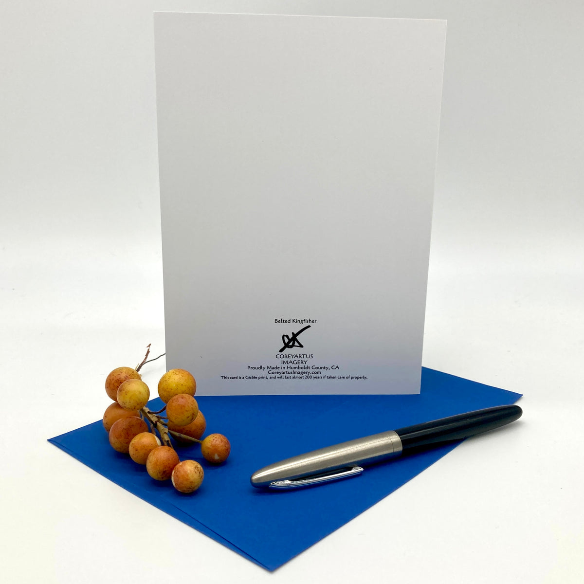 The back of a greeting card, posed with a sprig of yellow berries, an ink pen, and a dark blue envelope.