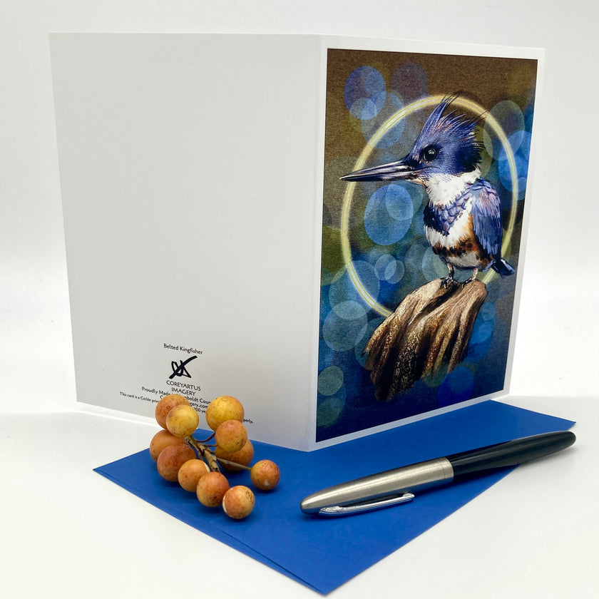 A splayed out, illustrated greeting card featuring a Belted Kingfisher bird on a blue background with abstract glowing bubble orbs behind it. Posed with a sprig of yellow berries, an ink pen, and a dark blue envelope.