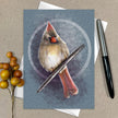 Card with a painted cardinal bird on a textured blue background, placed on a beige surface with a pen and decorative elements.