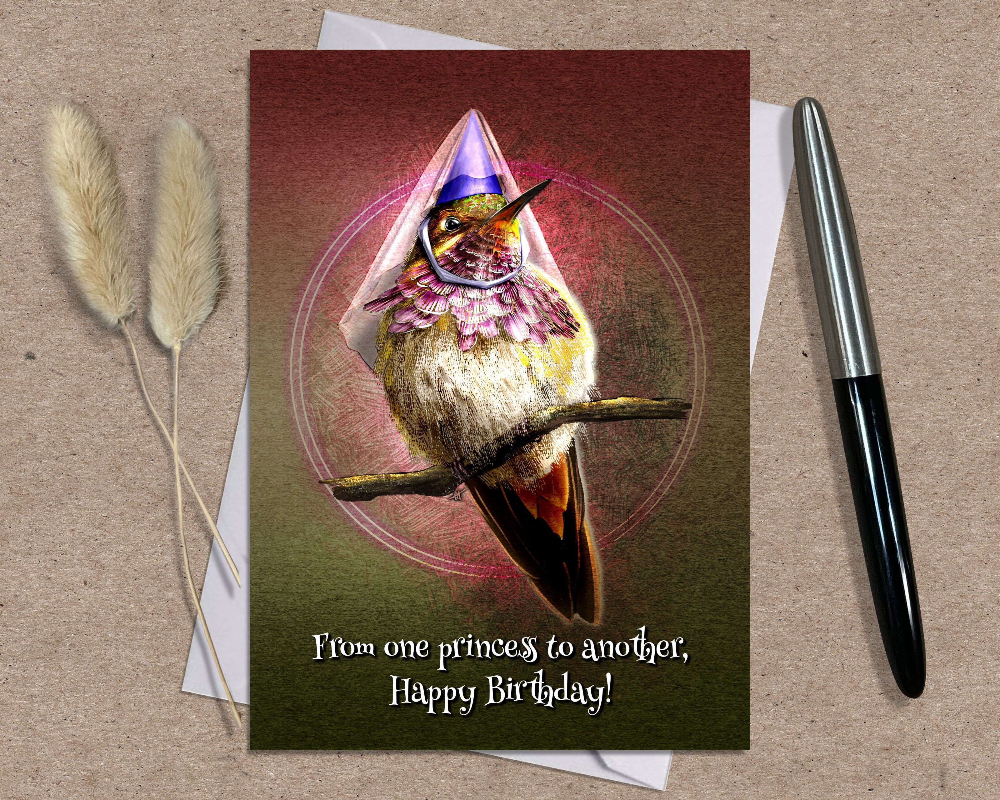 Greeting card with a hummingbird wearing a princess dress on a brown surface with a pen and dried plants.