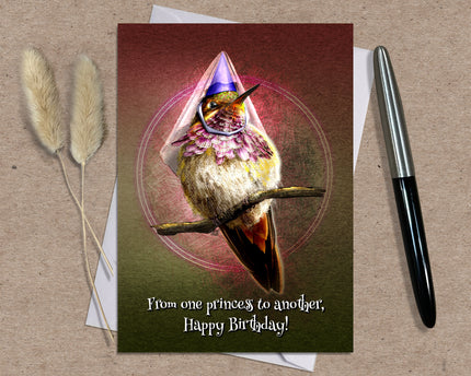 Greeting card with a hummingbird wearing a princess dress on a brown surface with a pen and dried plants.