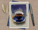 A greeting card featuring a watercolor-style illustration of a dark blue teacup and saucer with gold detailing, set against a textured beige background. The print is displayed flat on a brown kraft paper surface, layered over a blue card, with two dried pampas grass stems on the left and a black pen on the right.