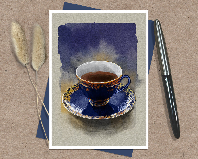 A greeting card featuring a watercolor-style illustration of a dark blue teacup and saucer with gold detailing, set against a textured beige background. The print is displayed flat on a brown kraft paper surface, layered over a blue card, with two dried pampas grass stems on the left and a black pen on the right.