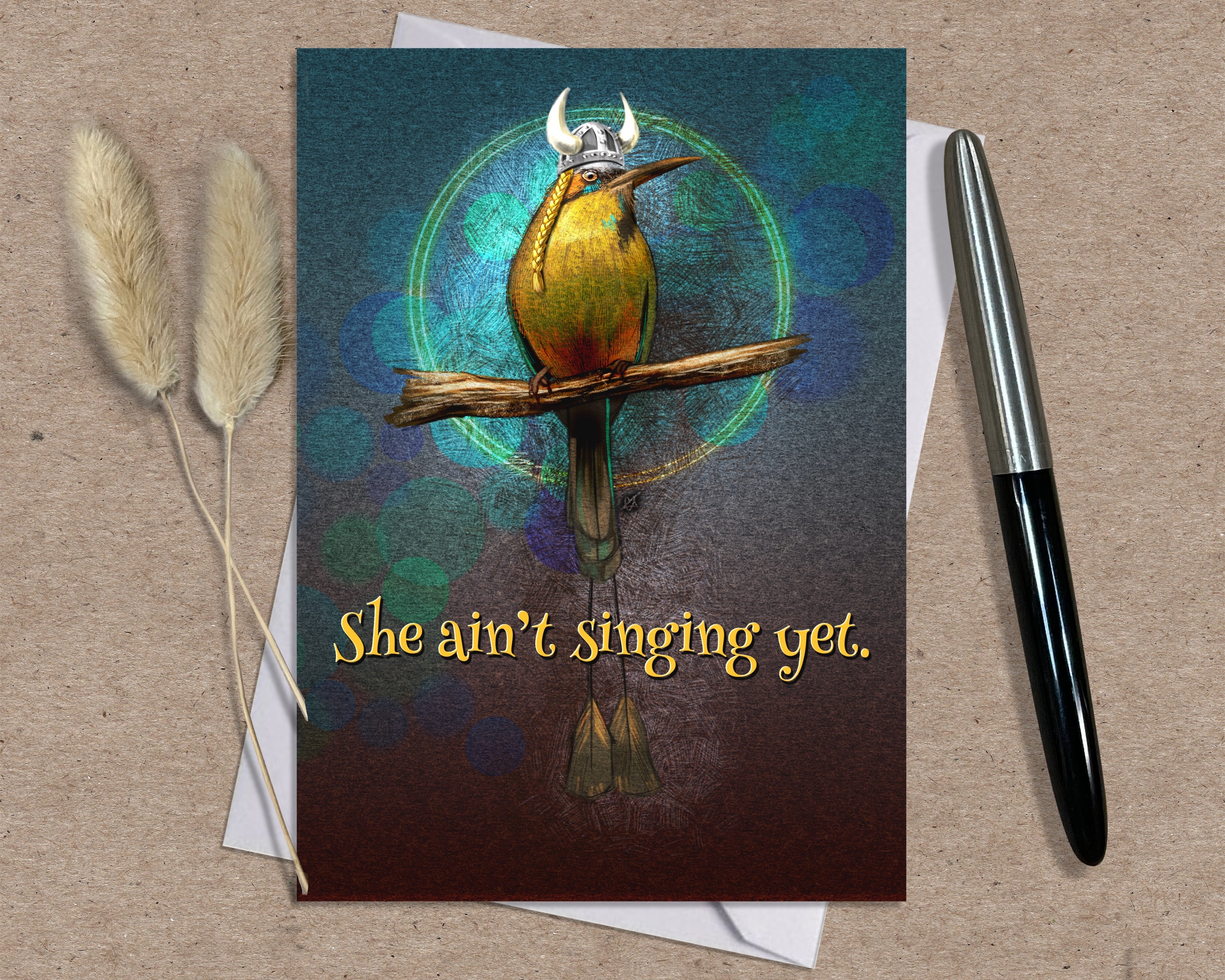 Greeting card with a bird illustration and text 'She ain't singing yet.' on a textured surface with a pen and dried plants.