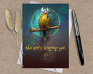 Greeting card with a bird illustration and text 'She ain't singing yet.' on a textured surface with a pen and dried plants.