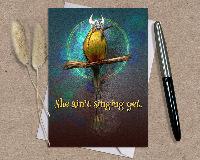 Greeting card with a bird illustration and text 'She ain't singing yet.' on a textured surface with a pen and dried plants.