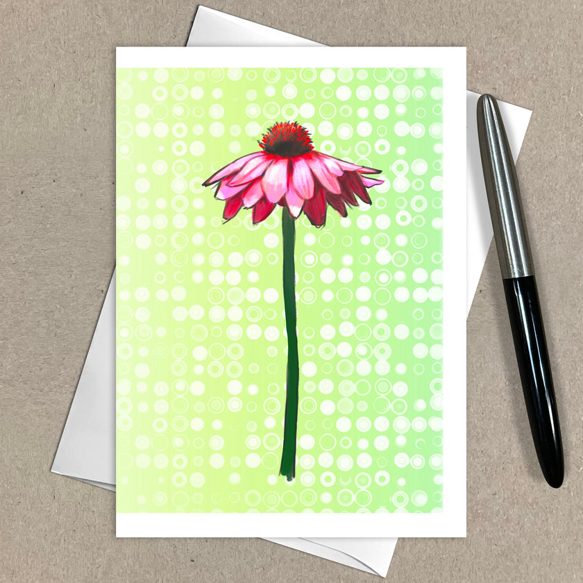 Greeting card with pink flower design on a green polka dot background, placed on a brown surface with a pen.