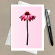 Greeting card with a pink flower illustration on a pink background, placed on a brown surface with a pen.