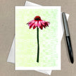 Greeting card with a pink flower design on a light green background, placed on a brown surface with an envelope and pen.