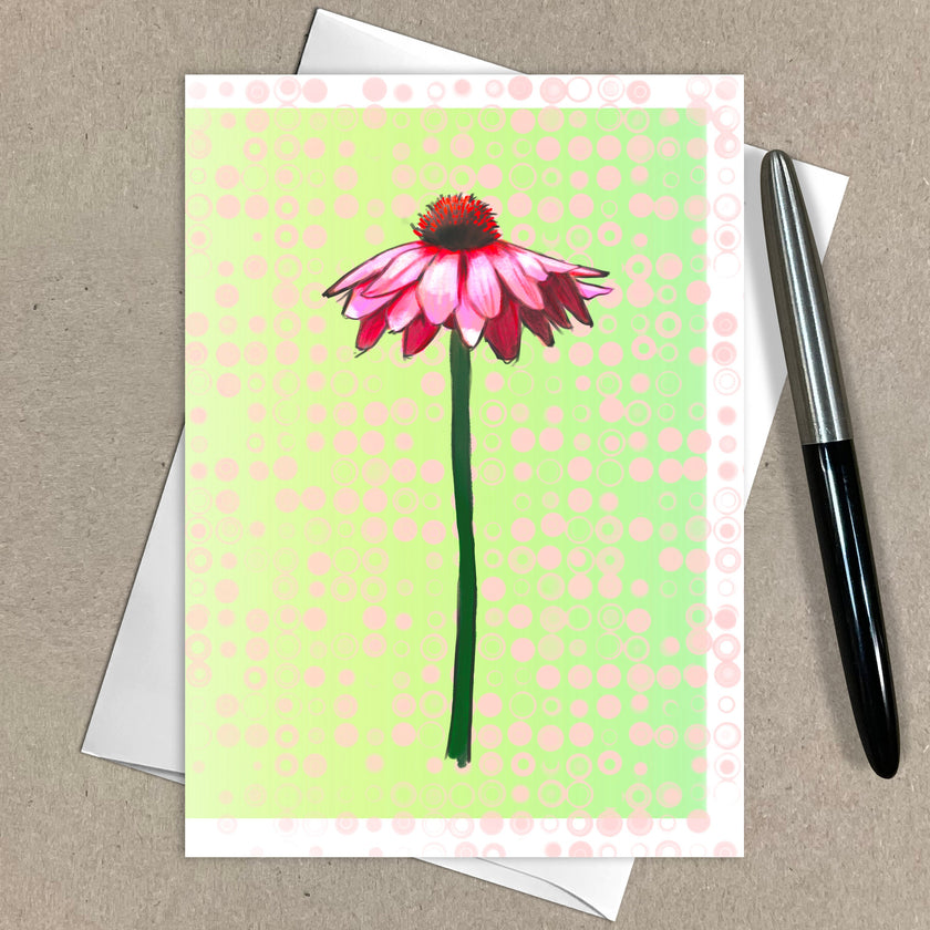 Greeting card with pink flower design on a green polka dot background, with an envelope and pen on a brown surface.