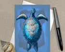 Greeting card with a blue turtle design on a textured surface with a pen and shell.