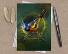 Greeting card with a colorful bird illustration on a textured surface with a pen and dried plants.