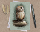 Greeting card with an owl illustration on a textured surface with a pen and dried plants.