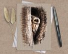 Greeting card with owl illustration on a textured surface with a pen and dried plants.