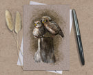 Greeting card with owl illustration on a textured surface with a pen and dried plants.