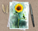 A square art print featuring a vibrant yellow sunflower with a textured teal and green watercolor background, styled on a brown kraft paper surface with a black pen and dried bunny tail grass.