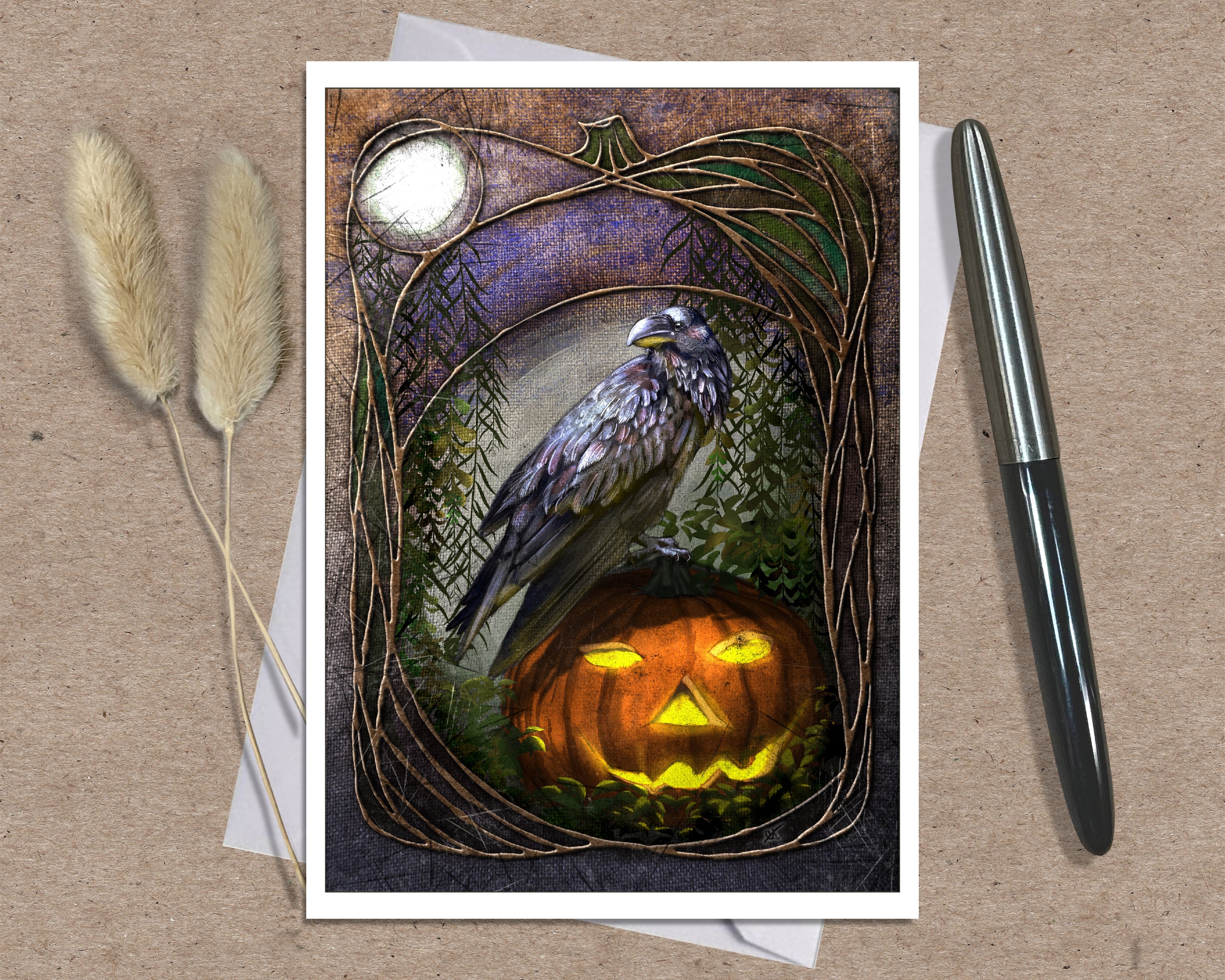 An art print greeting card featuring a raven perched on a glowing jack-o'-lantern, displayed on a kraft paper background with two dried grass stems and a black pen as props.