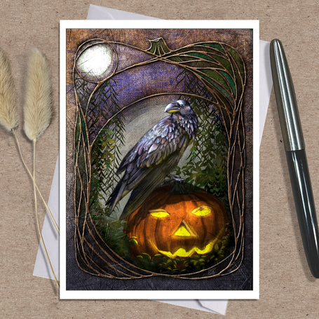An art print greeting card featuring a raven perched on a glowing jack-o'-lantern, displayed on a kraft paper background with two dried grass stems and a black pen as props.