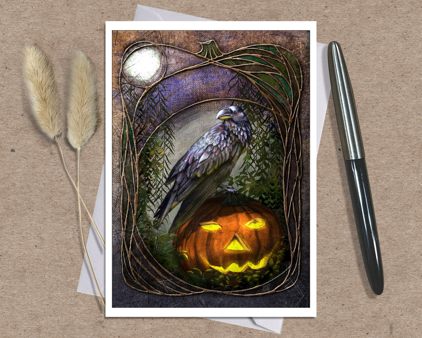 An art print greeting card featuring a raven perched on a glowing jack-o'-lantern, displayed on a kraft paper background with two dried grass stems and a black pen as props.