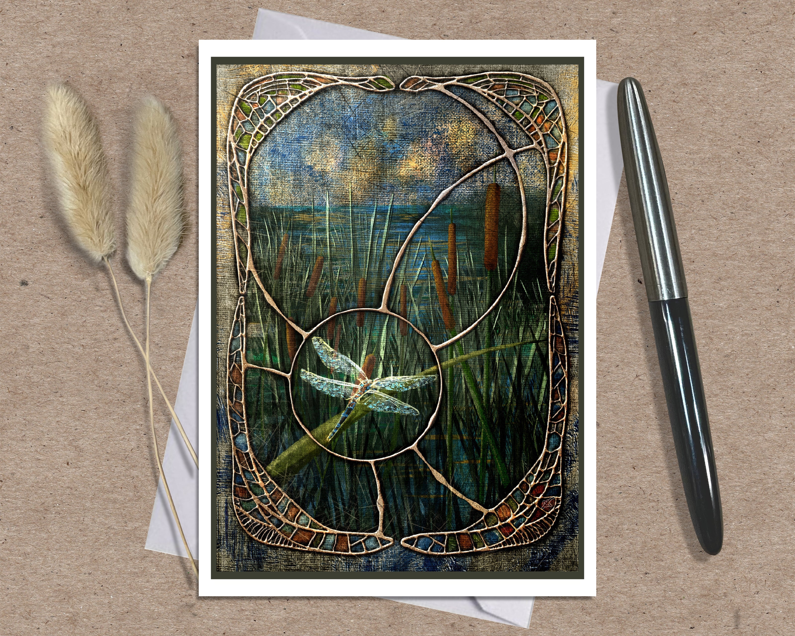 A square art print or card featuring a stained-glass-style reeds and dragonfly scene, displayed on a kraft paper background with two dried grass stems on the left and a black pen on the right.
