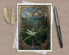 A square art print or card featuring a stained-glass-style reeds and dragonfly scene, displayed on a kraft paper background with two dried grass stems on the left and a black pen on the right.