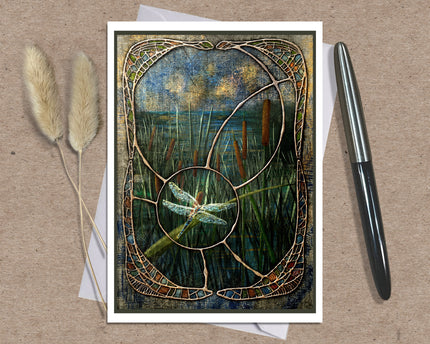 A square art print or card featuring a stained-glass-style reeds and dragonfly scene, displayed on a kraft paper background with two dried grass stems on the left and a black pen on the right.