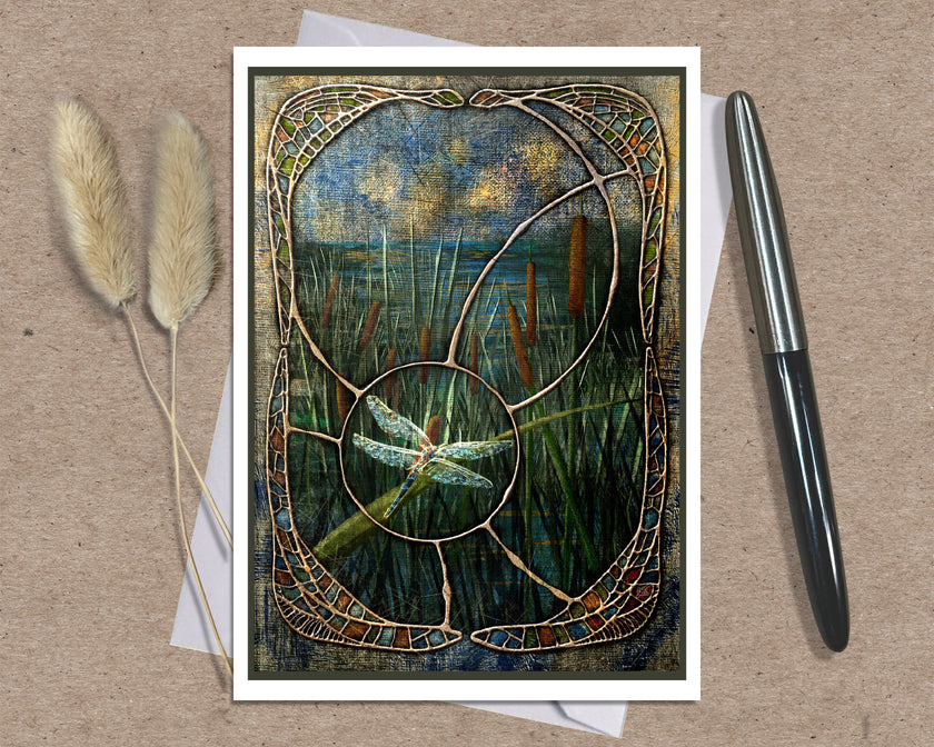 A square art print or card featuring a stained-glass-style reeds and dragonfly scene, displayed on a kraft paper background with two dried grass stems on the left and a black pen on the right.