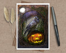 Product shot of an illustrated Halloween greeting card featuring a raven and a jack o'lantern, displayed on a brown envelope with a sprig of natural grasses and an ink pen as complimentary props.