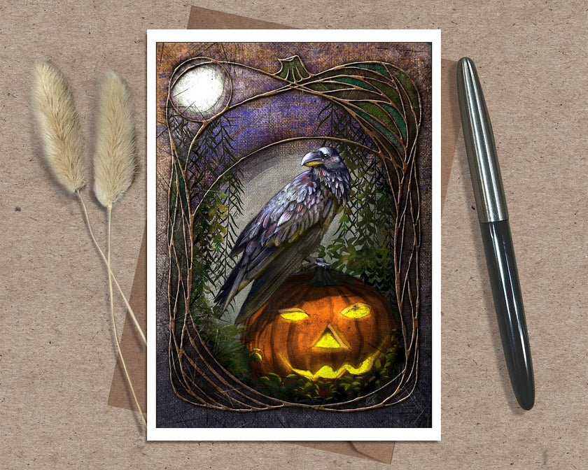 Product shot of an illustrated Halloween greeting card featuring a raven and a jack o'lantern, displayed on a brown envelope with a sprig of natural grasses and an ink pen as complimentary props.