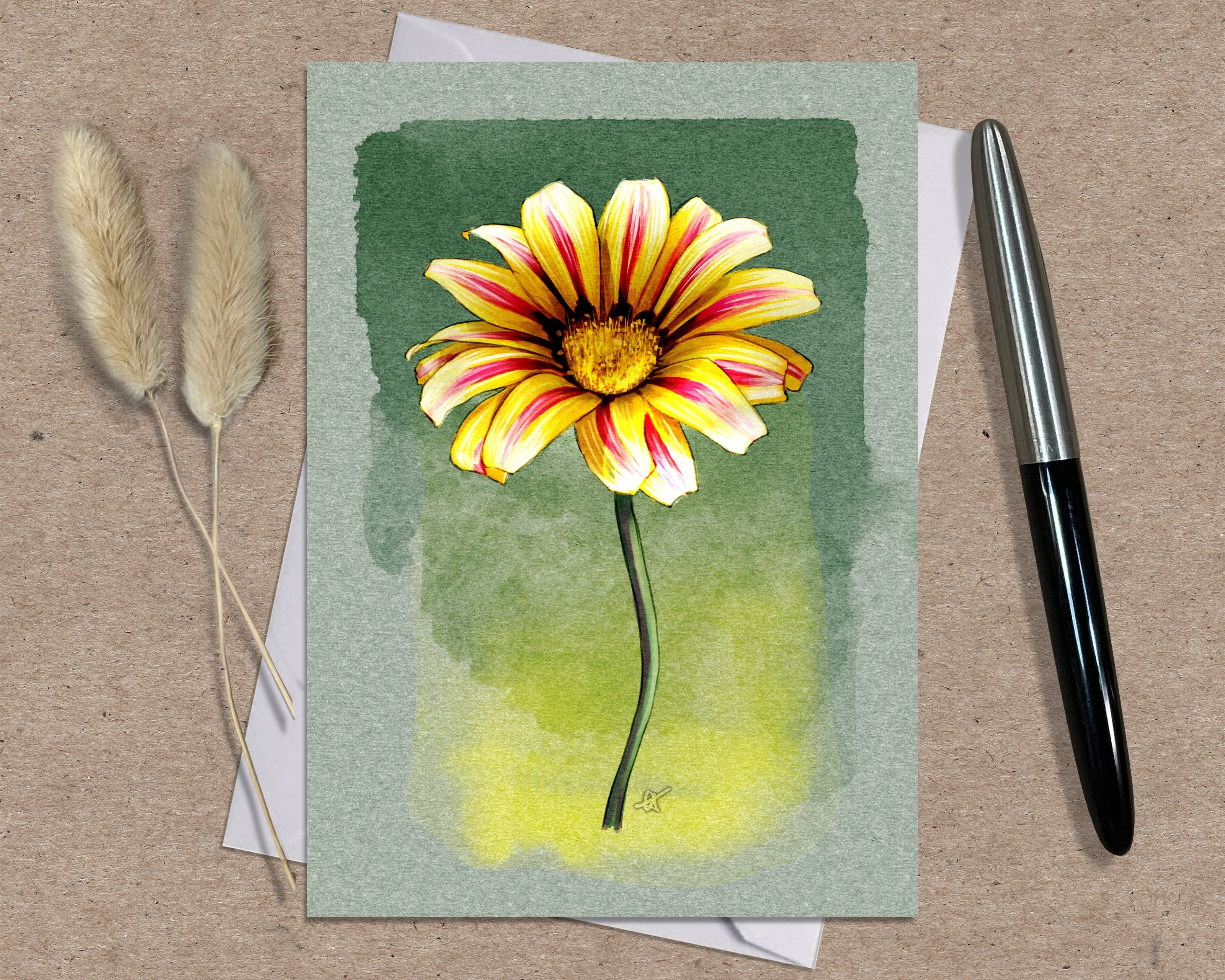 Greeting card with a yellow and pink flower illustration on a green background, placed on a textured surface with a pen and dried plants.