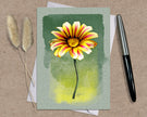 Greeting card with a yellow and pink flower illustration on a green background, placed on a textured surface with a pen and dried plants.