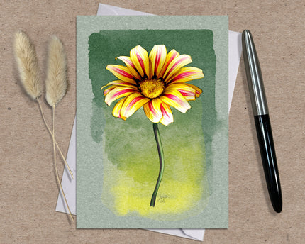 Greeting card with a yellow and pink flower illustration on a green background, placed on a textured surface with a pen and dried plants.