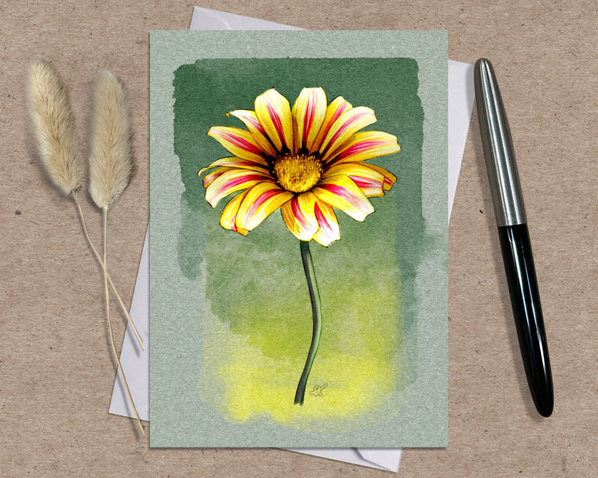 Greeting card with a yellow and pink flower illustration on a green background, placed on a textured surface with a pen and dried plants.